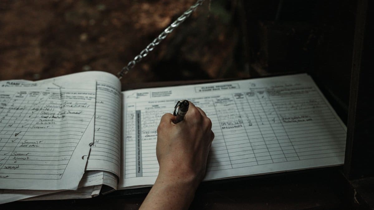 Close-up of a person writing in an outdoor logbook on a table.