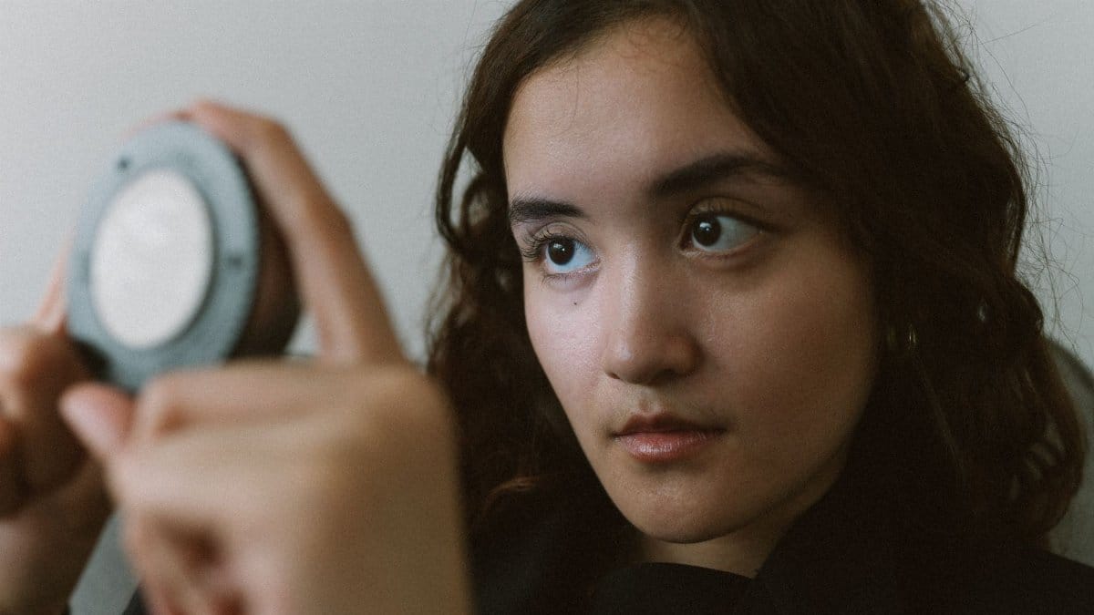 A young woman looking attentively at a compact mirror indoors, depicting self-reflection.