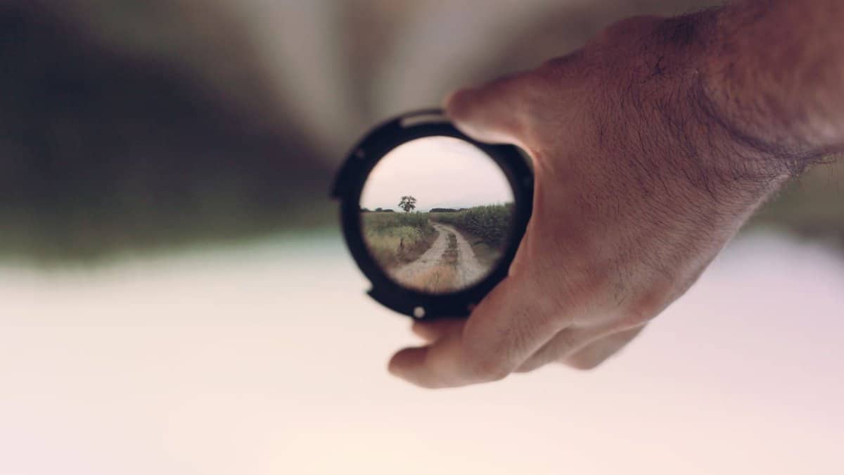 A hand holds a camera lens revealing a clear path through nature, symbolizing vision and focus.