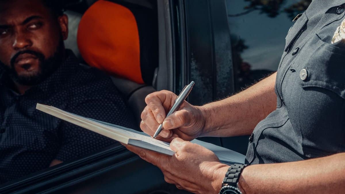 A police officer writes a ticket as the driver looks on from inside the car.