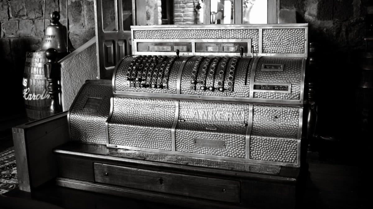 Black and white photo of a vintage Anker cash register in a rustic interior setting.