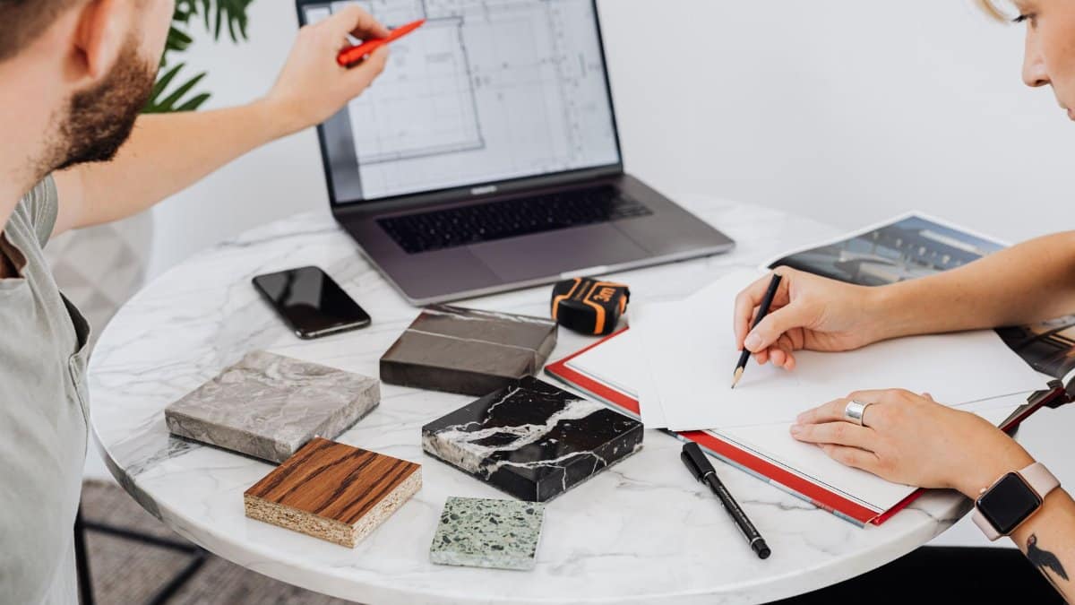 Two designers collaborate on a project with laptop and material samples on a marble table.