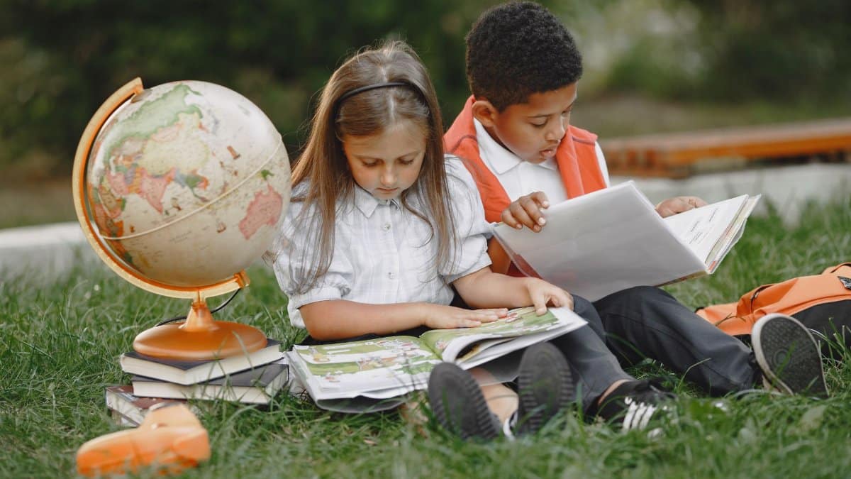 Two children sitting on grass reading books with a globe, promoting education.