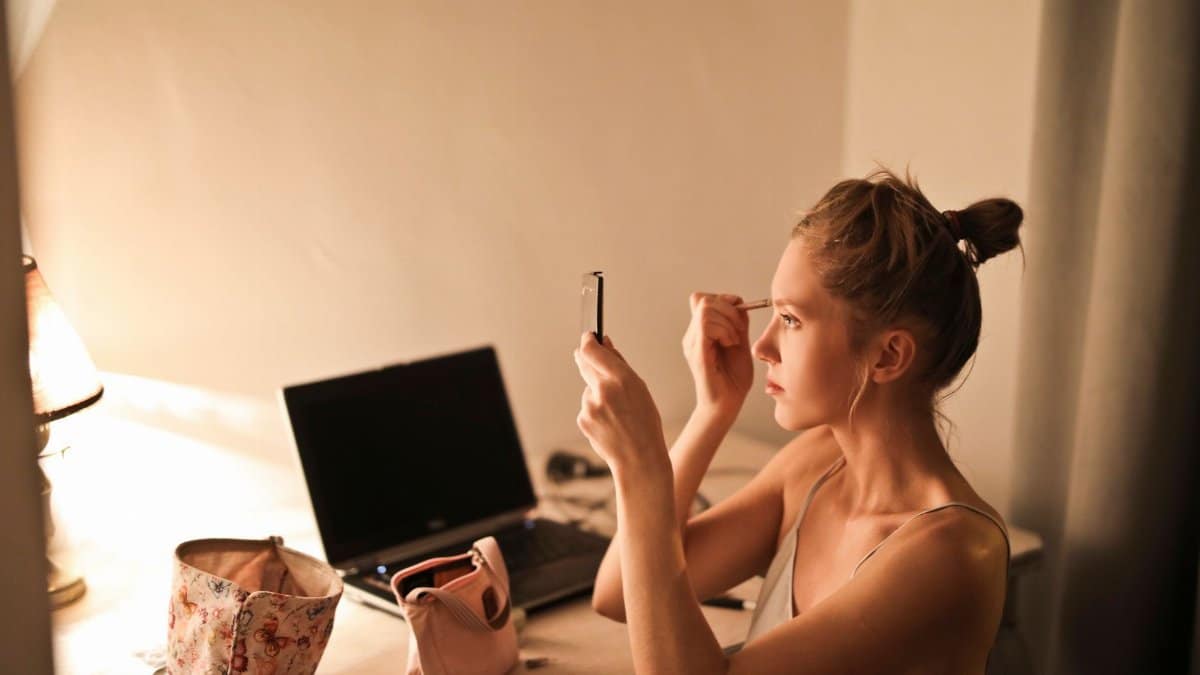 A young woman applies makeup at her vanity table, illuminated by soft light, in a cozy home setting.