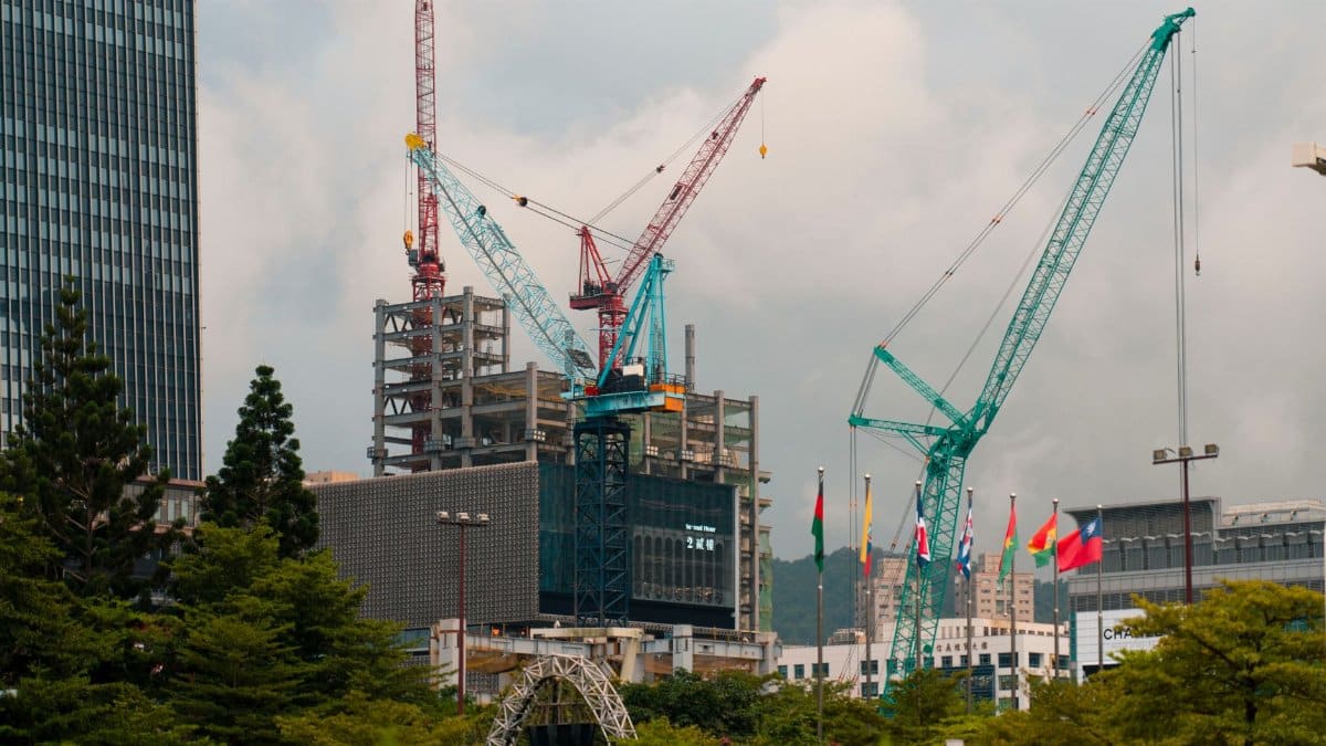 Cranes tower over a construction site amidst Taipei's urban landscape, featuring flags and a modern skyline.