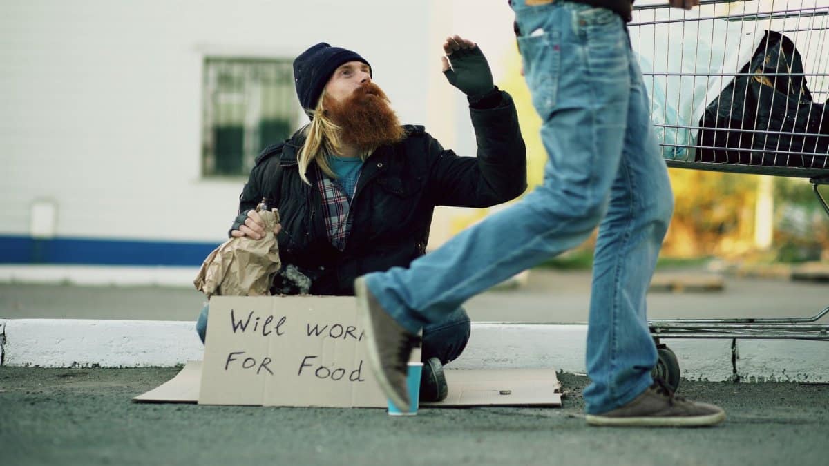 Homeless man sitting beside a shopping cart holding a sign reading 'Will Work For Food'.