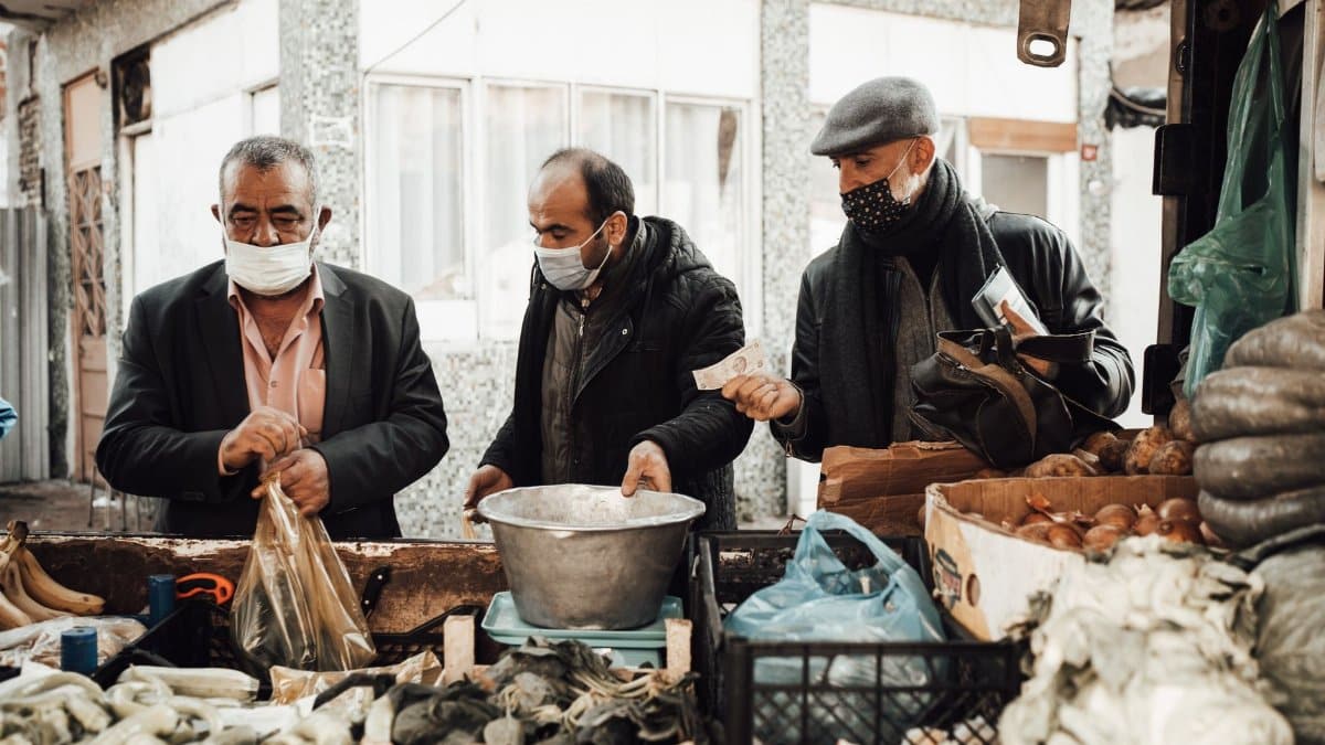 Multiethnic male buying food from seller in outerwear and protective mask near stall with different vegetables in boxes near metal basin and packets in local market in city street