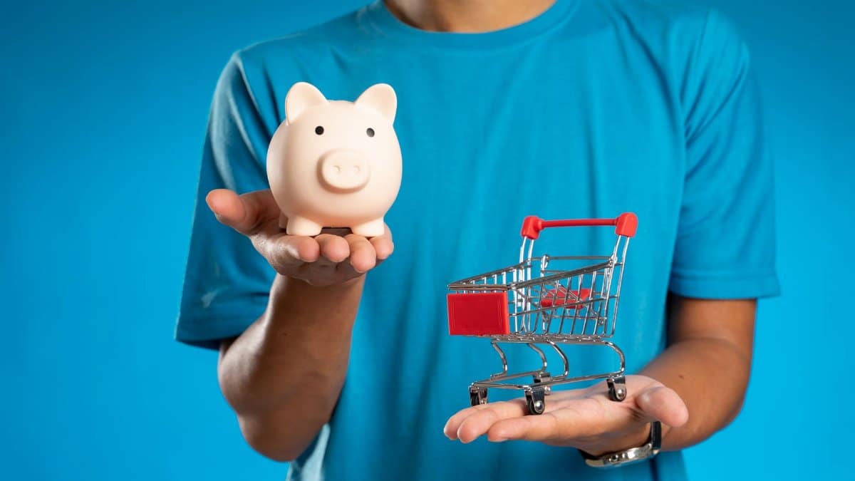 Man in blue shirt holding piggy bank and mini shopping cart, symbolizing savings versus spending.