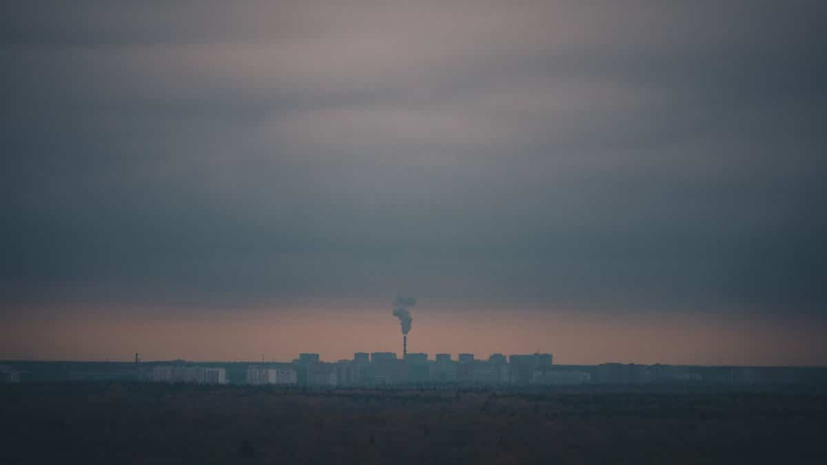 A distant view of a cityscape with a factory emitting smoke under a moody sky.