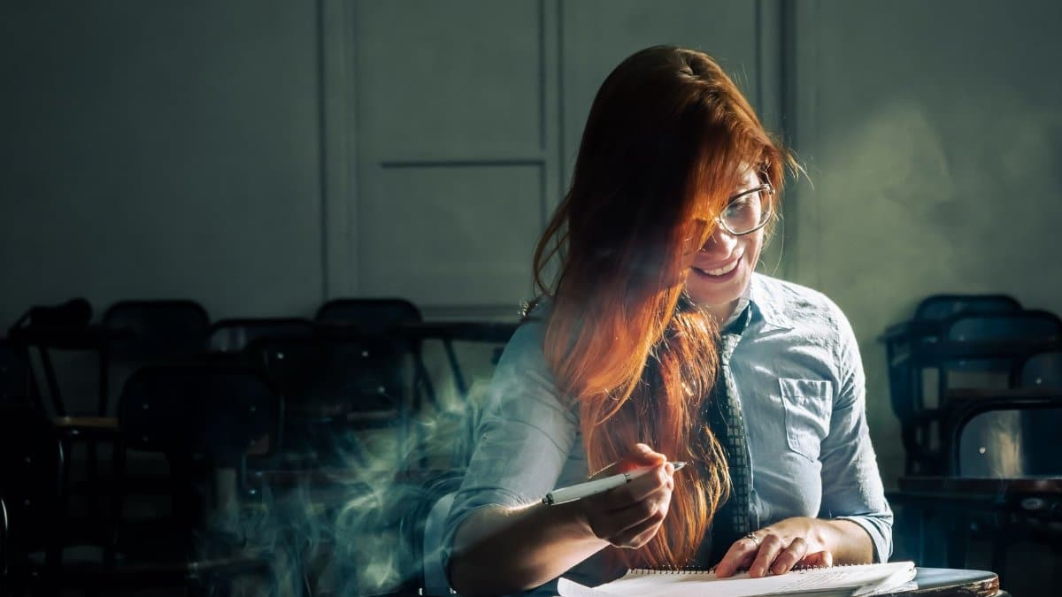 A young woman sits alone in a dimly lit classroom, writing and studying intently.