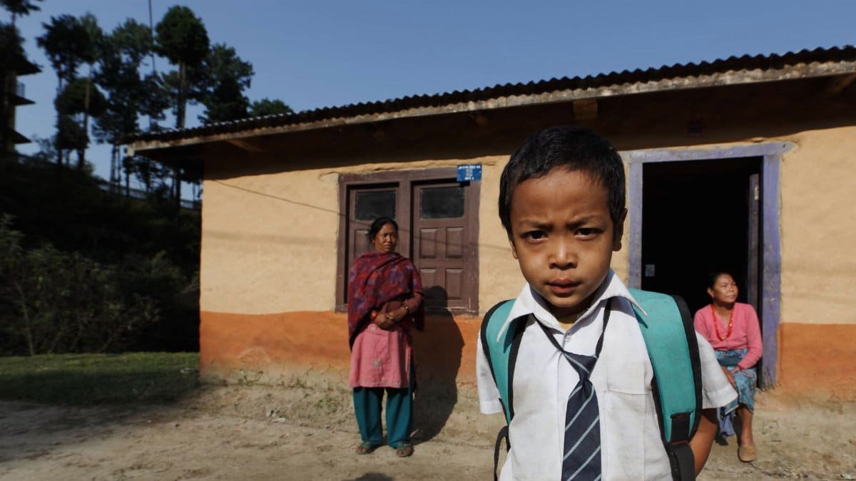 A young boy in school uniform stands in front of a rural house with two women in the background.