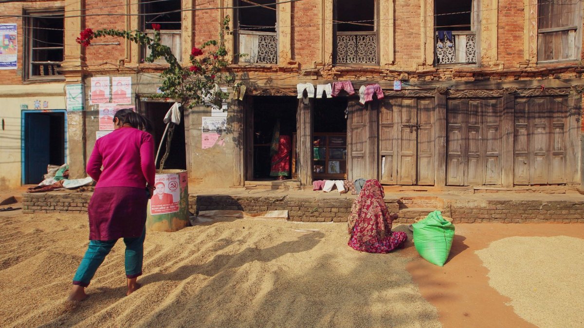 Two women drying grains outside a historic building in a rustic village.