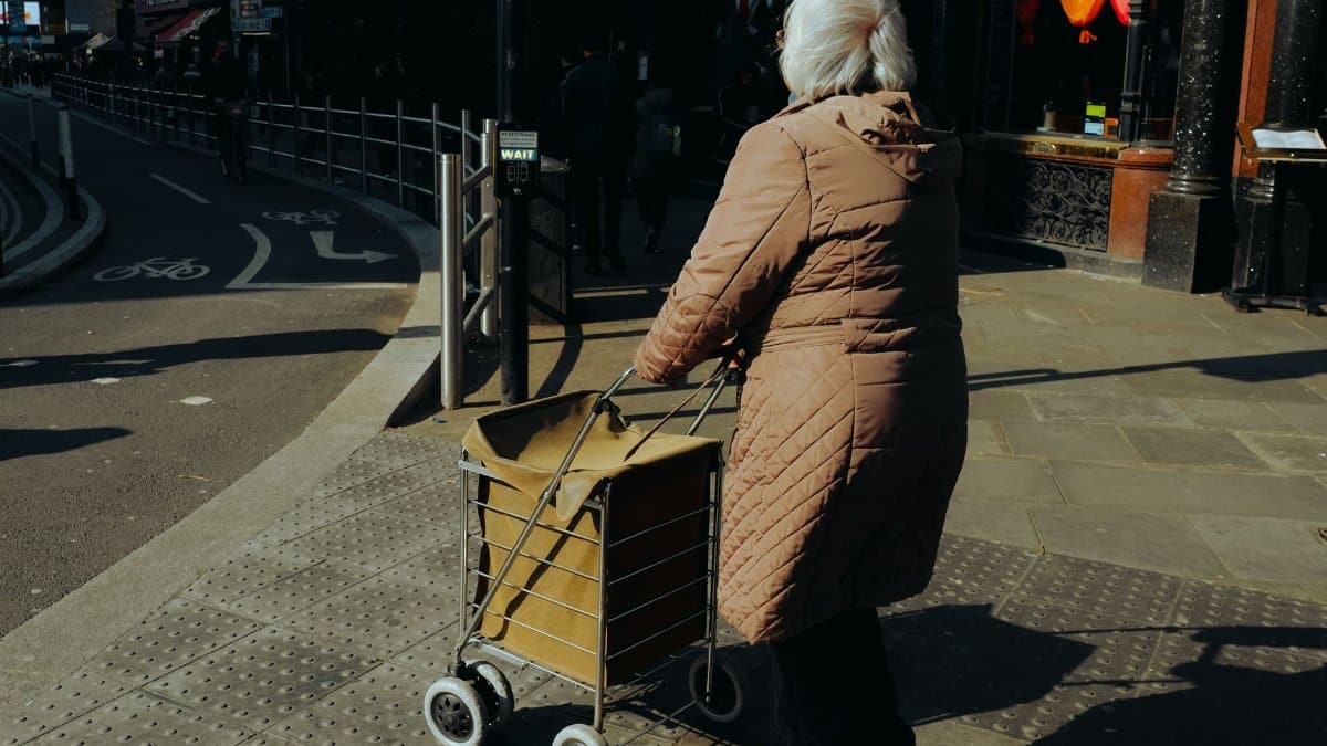 Elderly woman pushing a cart on a sunny city sidewalk, showcasing urban life.