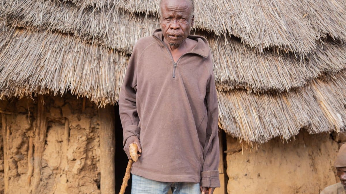 An elderly man stands in front of a rustic mud hut with a thatched roof, showcasing rural life.