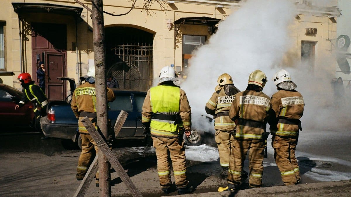 Firefighters responding to a car fire on a street in St. Petersburg, Russia, with smoke and safety gear in view.