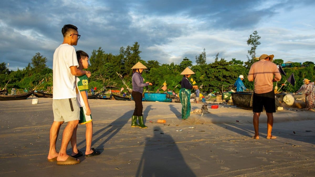 People on a beach engaging in traditional fishing during sunrise with boats in the background.