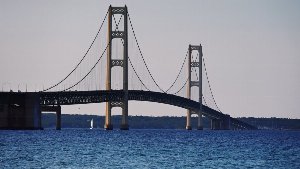 Serene view of Mackinac Bridge spanning the straits with a sailboat passing underneath.