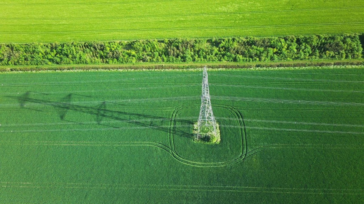 Aerial view of a transmission tower casting its shadow over lush green farmland, showcasing rural energy infrastructure.
