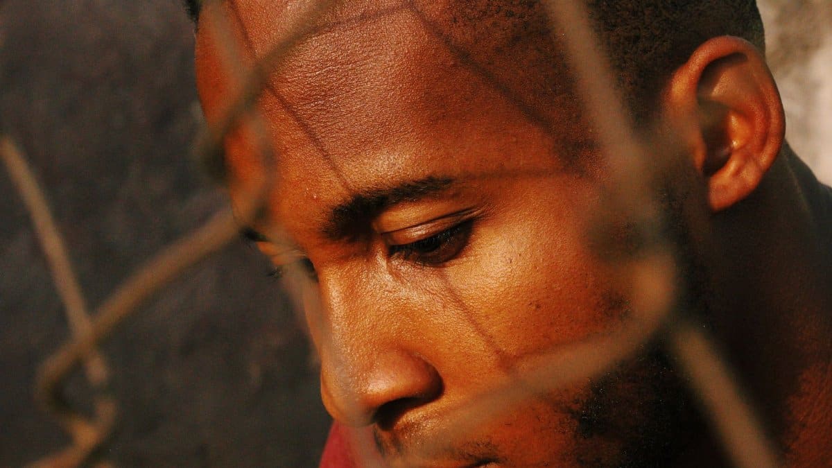 Close-up of a thoughtful young African man behind a rusted mesh fence in the sunlight.