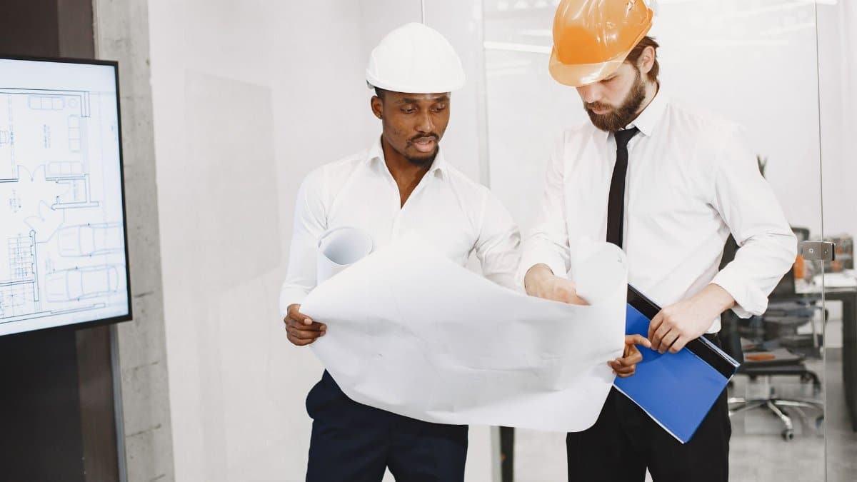 Two architects in hard hats review blueprints during a collaborative meeting in an office setting.