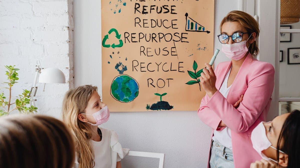 A teacher instructs masked students on environmental topics with a visual aid poster indoors.