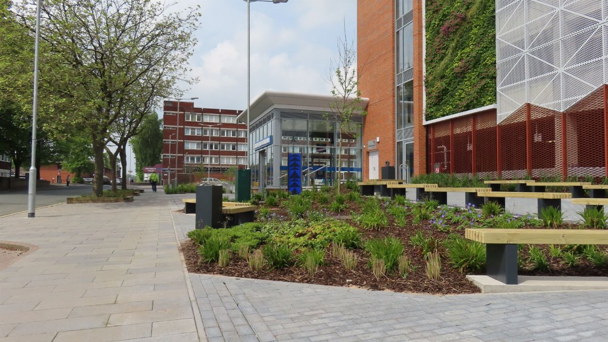 A modern urban street with a living wall on a building, benches, and well-kept greenery.