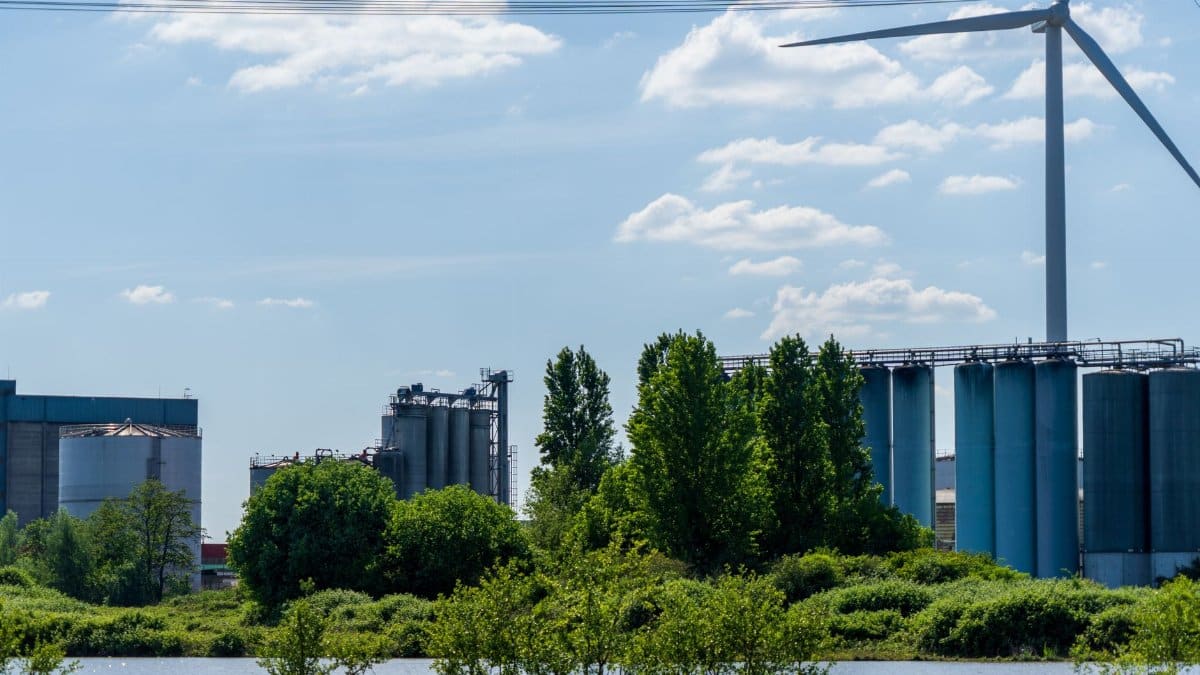 Scenic industrial landscape featuring a wind turbine, factories, and lush greenery by a body of water.
