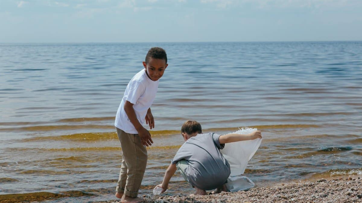Two children clean up a beach, collecting trash into a bag, promoting environmental awareness.