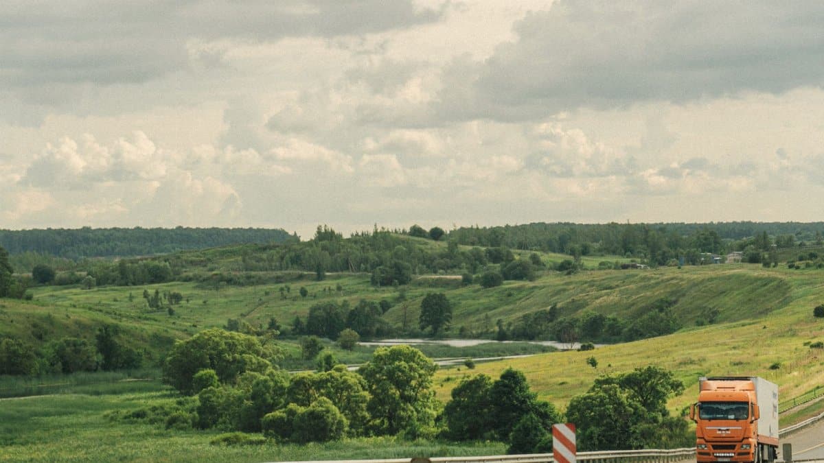 An orange truck drives along a highway through a lush, green, hilly countryside under a cloudy sky.