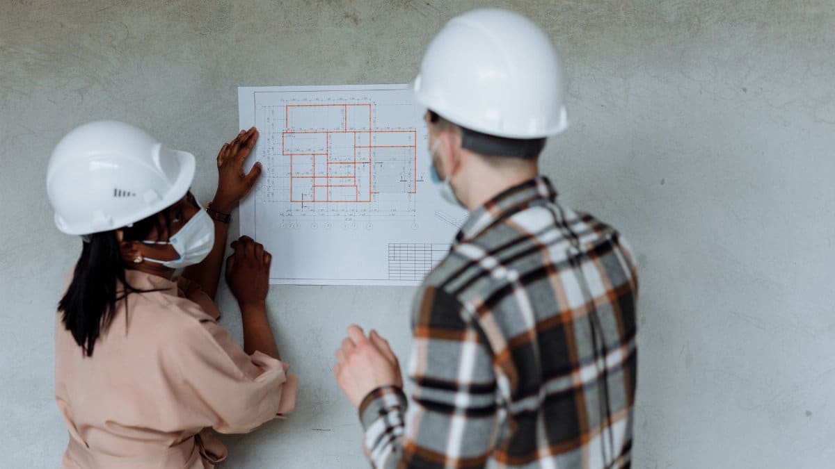 Two architects in hard hats and masks review blueprints during a building project.