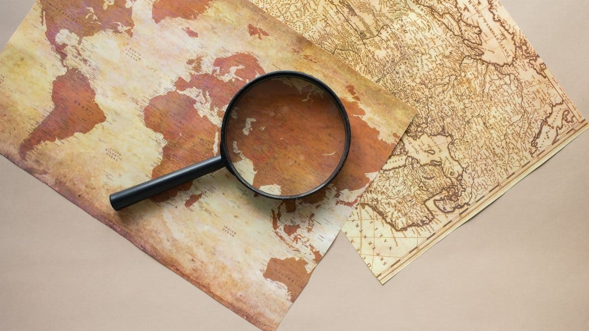 Top view of round black magnifying glass placed on paper maps with continents on beige background in light modern room