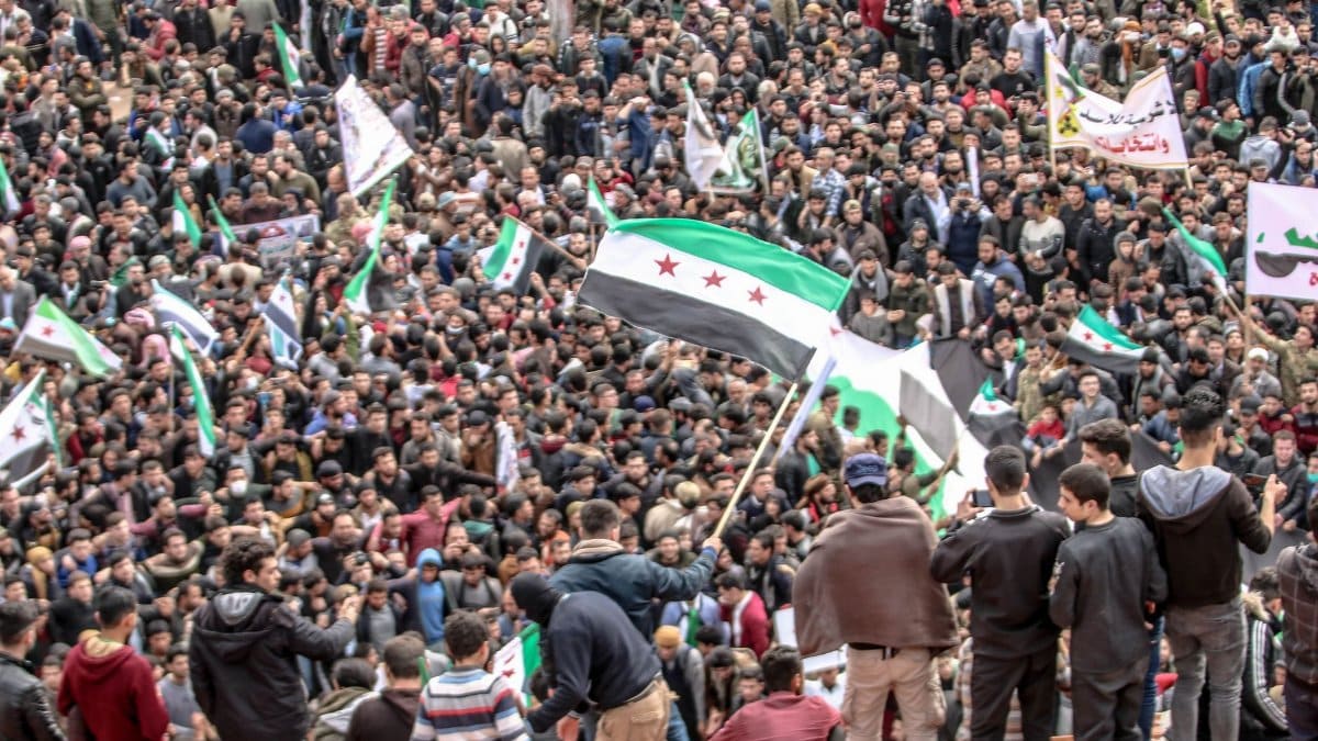 A large crowd protesting in Idlib, Syria, with flags and banners amid political unrest.