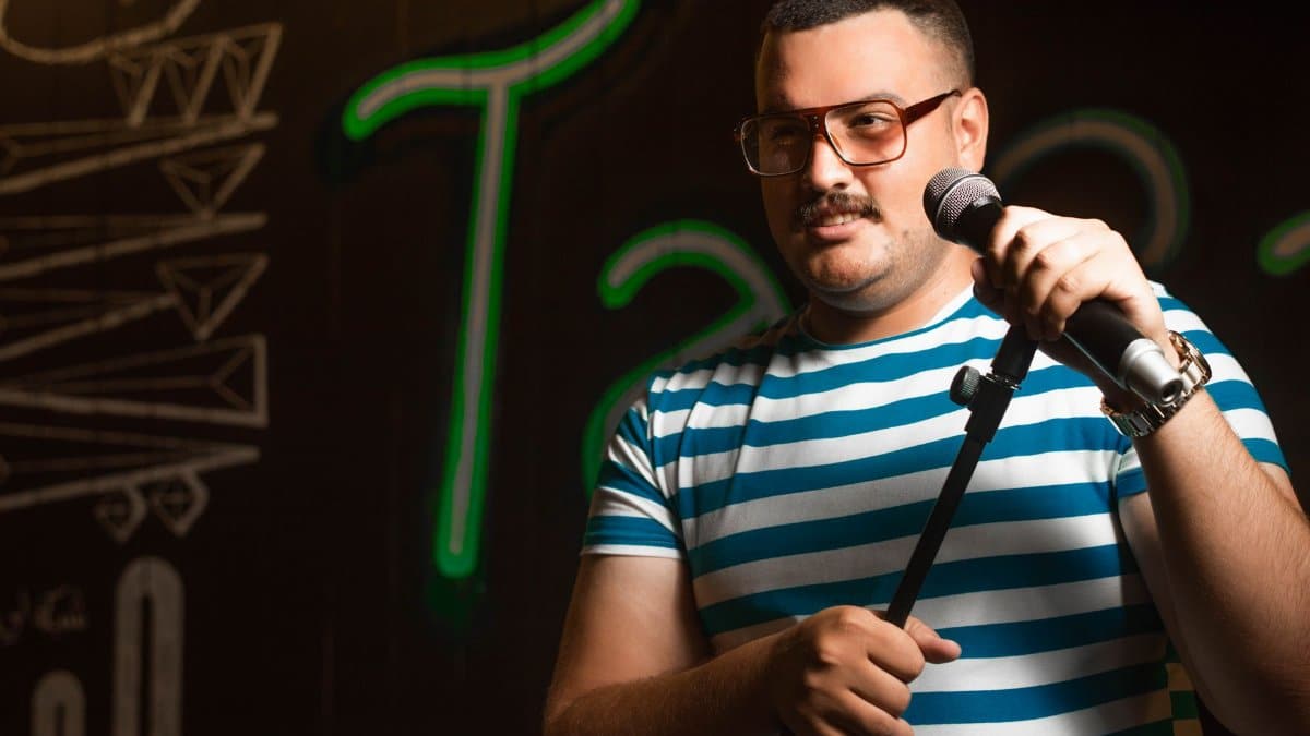 Man holding a microphone doing stand-up comedy in casual striped shirt on stage.