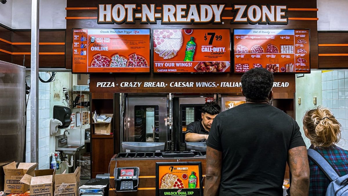 Busy fast food restaurant with customers ordering pizza at the counter under bright digital menu signage.