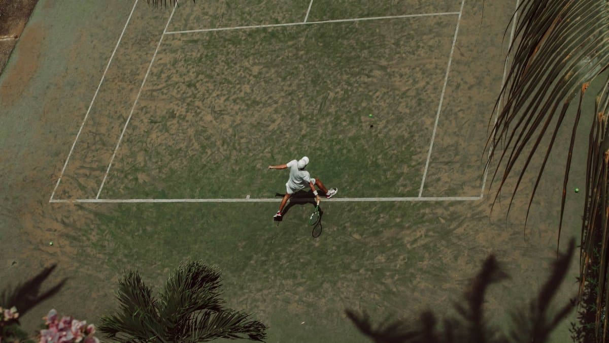 Aerial shot of a tennis player mid-action on a tropical court surrounded by lush vegetation.