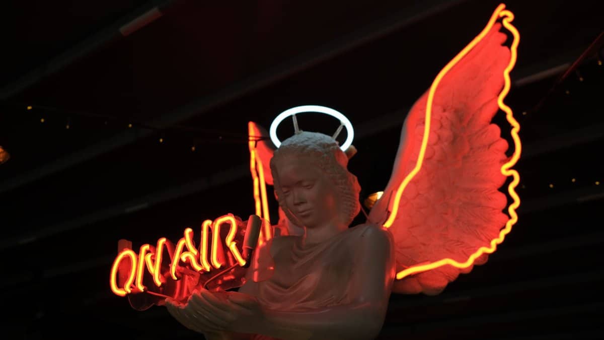 A statue of an angel with neon wings and halo, illuminated by a glowing 'On Air' sign.