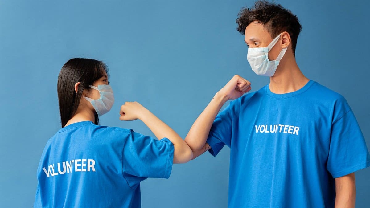 Two volunteers wearing blue shirts and face masks elbow bump for safety.