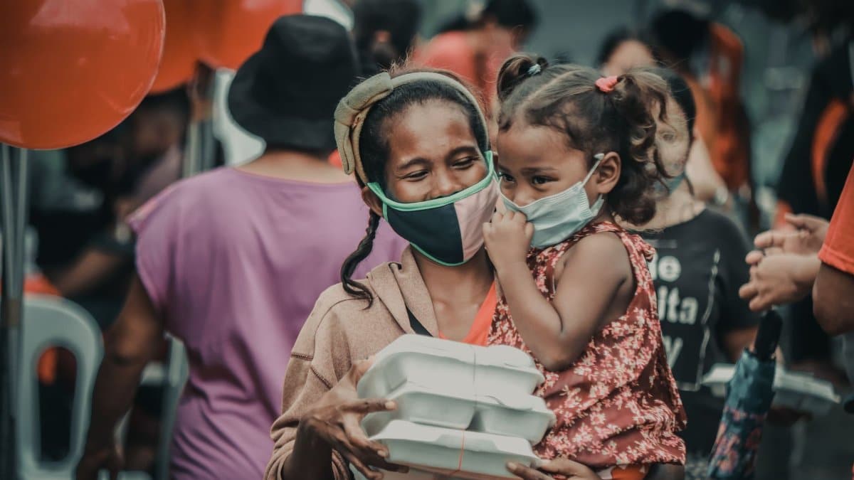 A mother and child wearing masks receive food packages at an outdoor event with balloons.