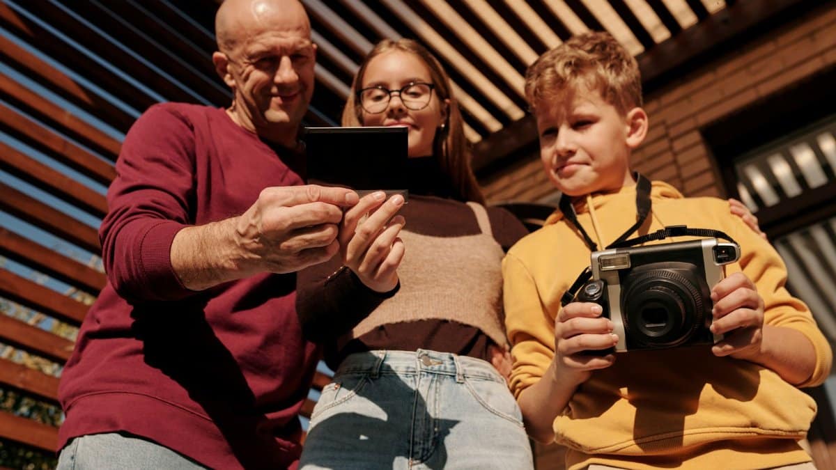 A family shares a joyful moment with a Polaroid camera under a pergola in a sunny outdoor setting.