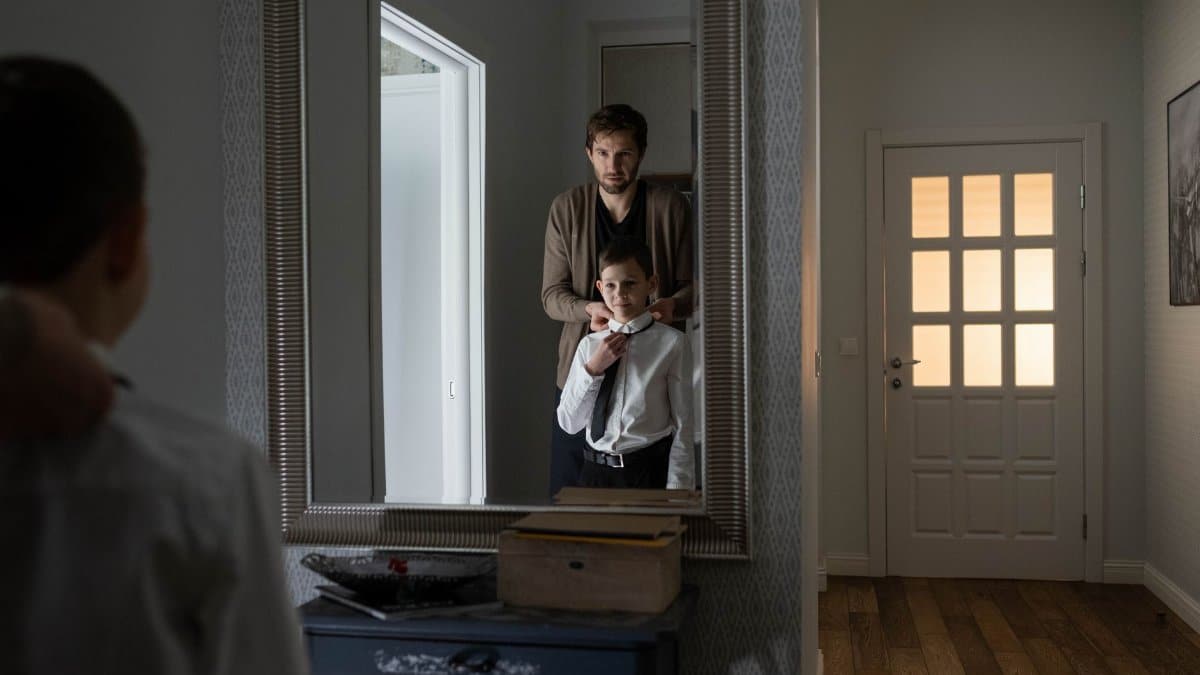 A father assists his son with tying a tie, reflected in a mirror, symbolizing family bonds.