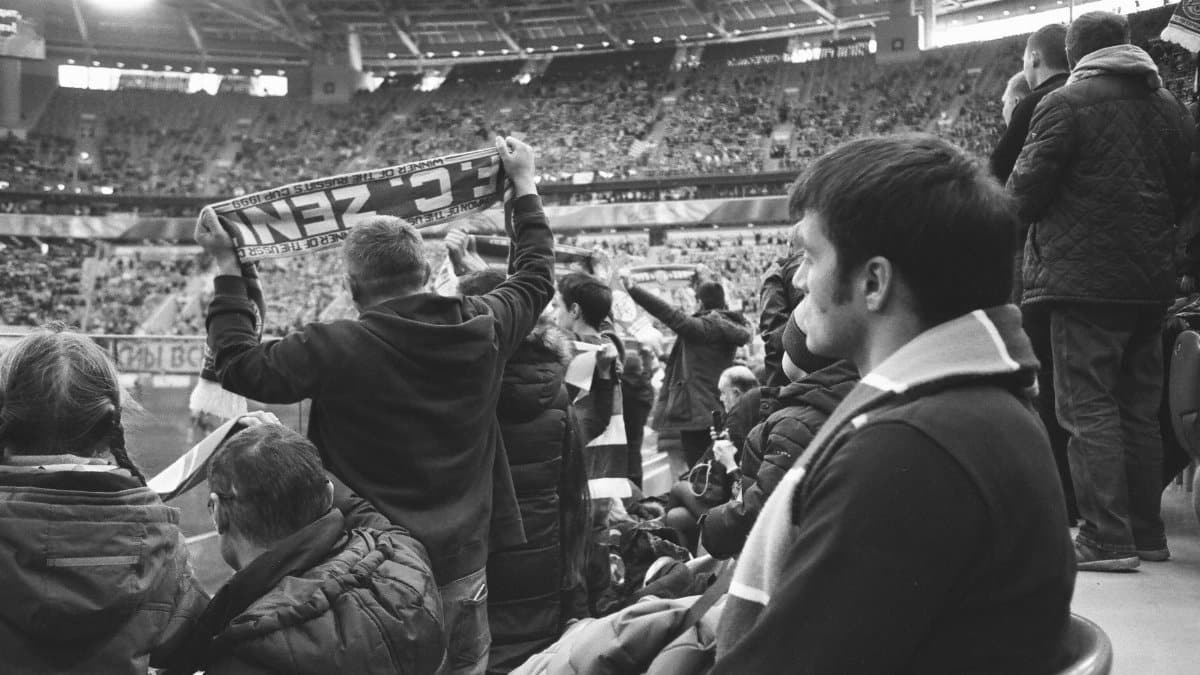 Black and white of group of people in warm clothing standing at stadium and watching match