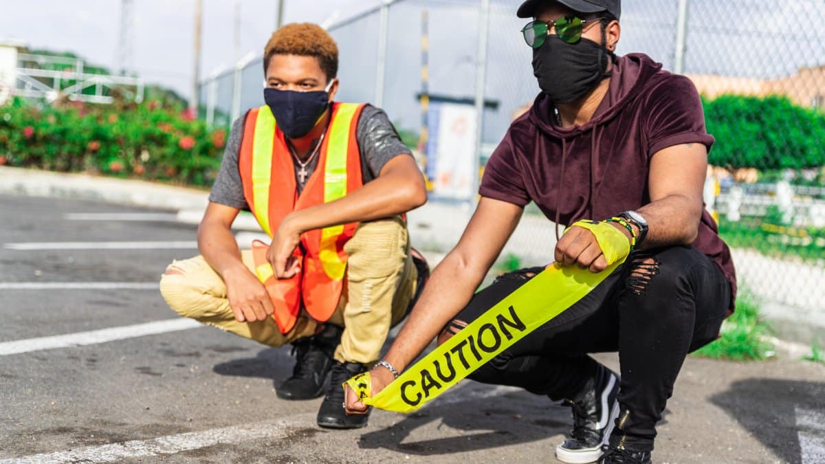 Two men in safety gear and masks crouching outdoors with caution tape.