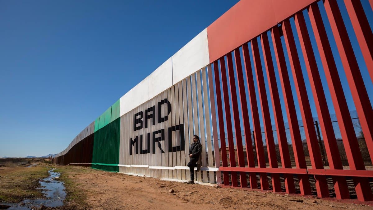 Image of a man standing by the border wall painted with Mexican flag colors in Naco, Sonora, Mexico.