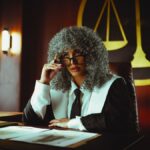 Judge in traditional attire sits at a courtroom desk with a serious expression.