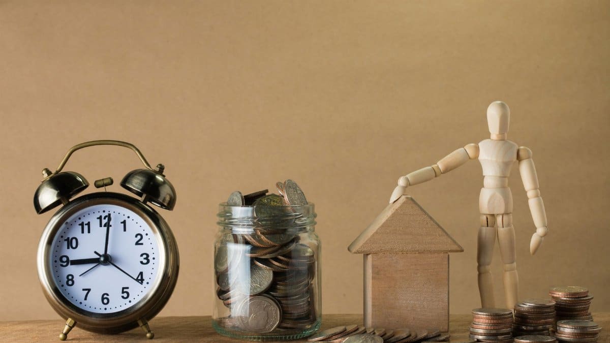 Wooden mannequin with a house, coins, and clock symbolizing time and financial planning.