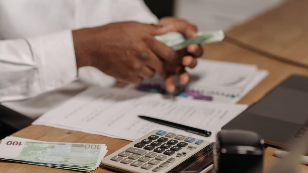 Hands counting euro bills on a wooden desk with calculator, financial documents, and laptop nearby.