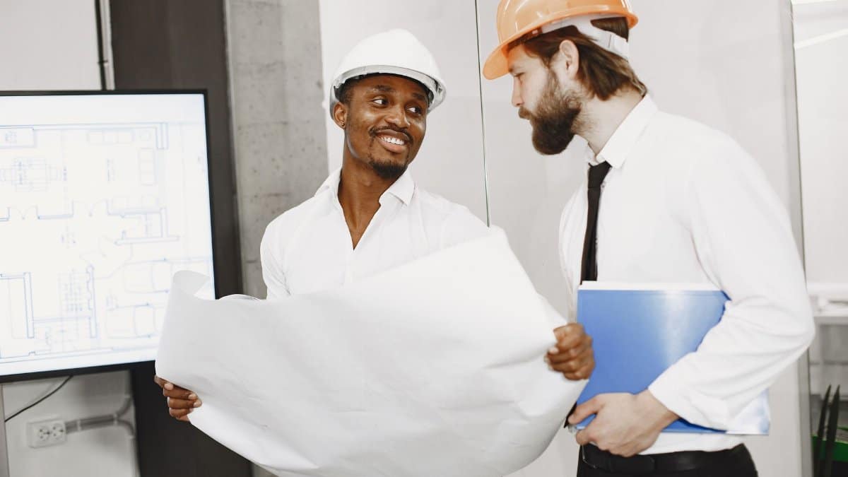 Two architects in hard hats discuss blueprints in a modern office setting, highlighting teamwork and planning.