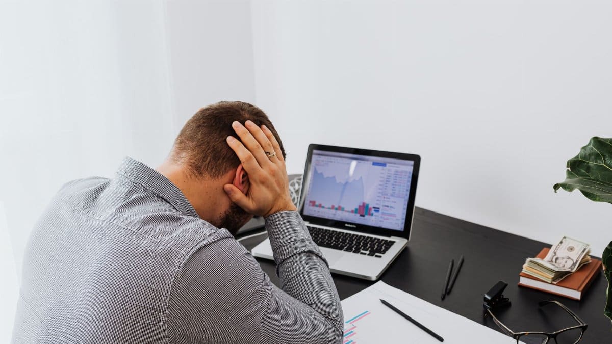 A businessman holds his head in frustration while sitting at a desk with a laptop and financial charts.