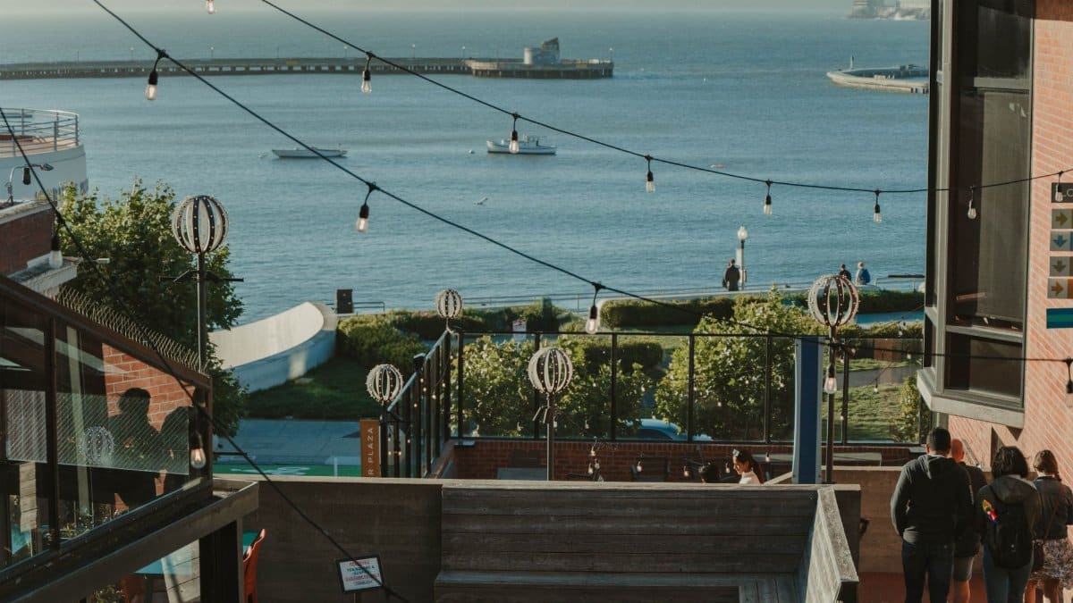 Terrace view of ocean bay with string lights, people, and boats at sunset; a serene outdoor scene.