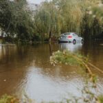 A car struggles through a flooded street surrounded by trees after a heavy rainstorm.